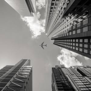 A low angle greyscale shot of an airplane flying above high rise buildings